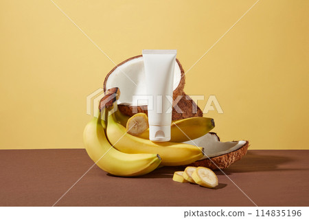 A unbranded tube of cosmetic displayed on a pile of fresh bananas and tropical coconut in the center of brown table over yellow background. Mockup scene for advertising with blank space for text A unbranded tube of cosmetic displayed on a pile of fresh bananas and tropical coconut in the center of brown table over yellow background. Mockup scene for advertising with blank space for text 114835196