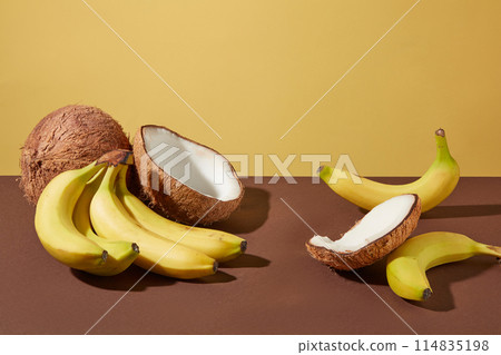 Tropical fruit including banana and coconut flat lay on brown table top, on yellow color texture. Vacant space in center for displaying cosmetic or beverage product manufactured from fruit ingredient Tropical fruit including banana and coconut flat lay on brown table top, on yellow color texture. Vacant space in center for displaying cosmetic or beverage product manufactured from fruit ingredient 114835198