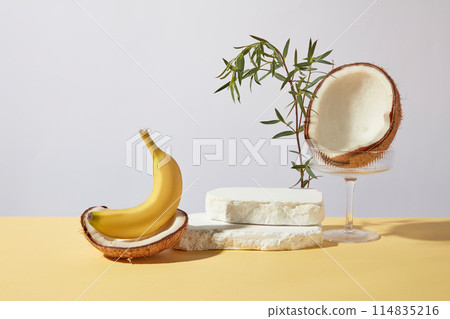 Creative photography with light blue background, two white plaster pedestal placed on top of each other into a higher podium, decorated with a glass, brown color of coconut and yellow color banana Creative photography with light blue background, two white plaster pedestal placed on top of each other into a higher podium, decorated with a glass, brown color of coconut and yellow color banana 114835216