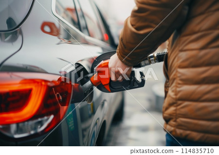 At a gas station, a person in a yellow jacket is fueling up a car using a fuel nozzle At a gas station, a person in a yellow jacket is fueling up a car using a fuel nozzle 114835710