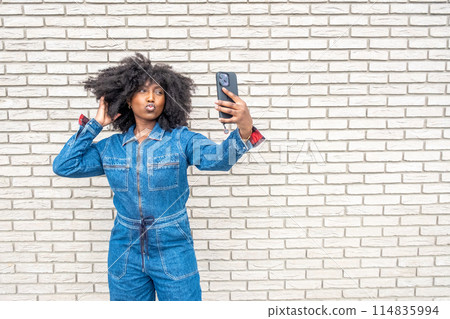 A woman in a denim jumpsuit taking a selfie against a brick wall backdrop A woman in a denim jumpsuit taking a selfie against a brick wall backdrop 114835994