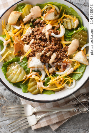 American salad with romaine lettuce, ground beef, pickles, cheddar cheese and croutons with sesame seeds close-up in a bowl on the table. Vertical top view 114838048