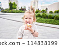 A hungry child enjoys eating ice cream outdoors. Portrait of a cute baby licking a creamy ice cream while walking in a city park, outdoor activities. 114838199