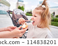 A hungry child enjoys eating ice cream outdoors. Portrait of a cute baby licking a creamy ice cream while walking in a city park, outdoor activities. 114838201