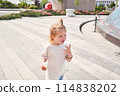 A hungry child enjoys eating ice cream outdoors. Portrait of a cute baby licking a creamy ice cream while walking in a city park, outdoor activities. 114838202