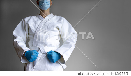 COVID-19 Pandemic Coronavirus concept. A young strong man in a white kimono for sambo, jiu jitsu and other martial arts with a blue medical gloves and medical mask. Grey background. Copy space. 114839585