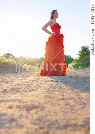 A woman in red dress poses gracefully on a rural road at sunset, creating a serene scene A woman in red dress poses gracefully on a rural road at sunset, creating a serene scene 114843859