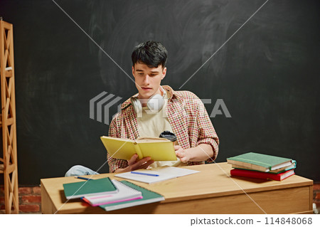 Young man, student sitting at desk and involved on book reading. Chalkboard background. Sitting in classroom and studying Young man, student sitting at desk and involved on book reading. Chalkboard background. Sitting in classroom and studying 114848068