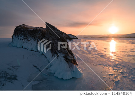 Aerial over the rocky island in lake Baikal. Winter landscape of frozen Baikal at beautiful orange sunrise. Sun reflections on the ice. Popular tourist spot 114848164