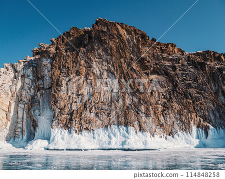 A majestic rocky cliff towers over the frozen surface of Lake Baikal, adorned with stunning ice formations and illuminated under a clear blue sky. A majestic rocky cliff towers over the frozen surface of Lake Baikal, adorned with stunning ice formations and illuminated under a clear blue sky. 114848258