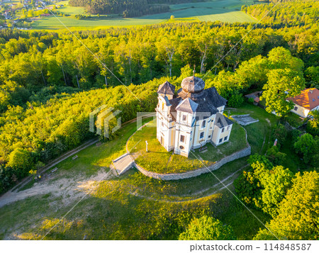 An aerial view of the Church of Saint John the Baptist and Our Lady of Mount Carmel at Makova hora, Czechia, surrounded by lush greenery on a bright spring evening. 114848587