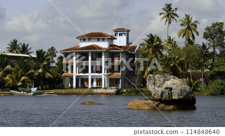 The view of mansion on the shore with palm trees. Action. Living at the sea shore with green vegetation. The view of mansion on the shore with palm trees. Action. Living at the sea shore with green vegetation. 114848640