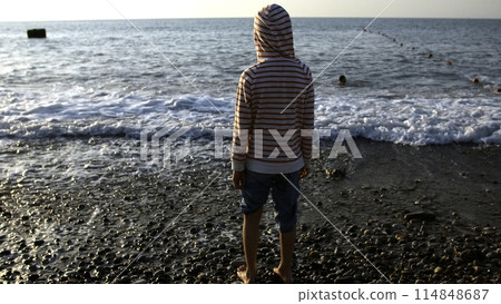 Happy child enjoys the sea on a hot summer day. Creative. Strong waves and boy standing barefoot on stony shore. Happy child enjoys the sea on a hot summer day. Creative. Strong waves and boy standing barefoot on stony shore. 114848687