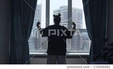 Rear view of a girl in casual clothes in modern bedroom in the morning. Media. Woman looking through the window, raising hands and stretching body muscles. Rear view of a girl in casual clothes in modern bedroom in the morning. Media. Woman looking through the window, raising hands and stretching body muscles. 114848802