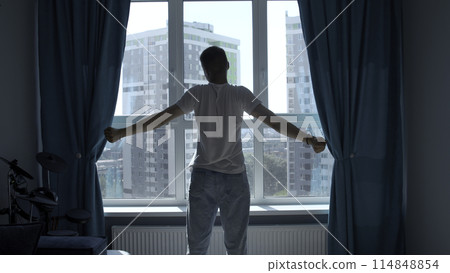 Early morning, silhouette of man exercising, stretching body by window at home. Media. Rear view of a young man standing against window. Early morning, silhouette of man exercising, stretching body by window at home. Media. Rear view of a young man standing against window. 114848854