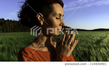 Young pretty girl woman meditating with closed eyes. Stock clip. Praying while standing in a field with a beautiful view on a windy summer day at sunset. 114848901