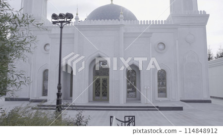 A white and gray historical mosque behind trees in the city. Scene. Religious building with minarets on cloudy sky background. 114848912