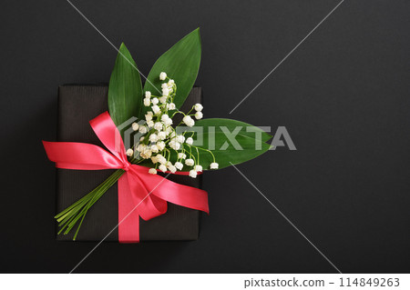 Black gift box with lilies of the valley and pink ribbon on dark background. Space for your text Black gift box with lilies of the valley and pink ribbon on dark background. Space for your text 114849263