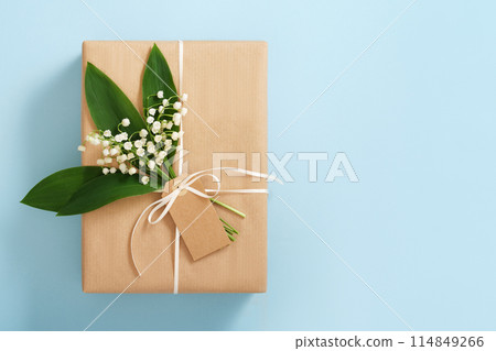 Card with brown gift box with empty rectangular tag and lilies of the valley on blue background. Space for your text 114849266