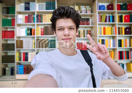 Selfie portrait of college student guy showing hand gesture for victory, inside library 114853080
