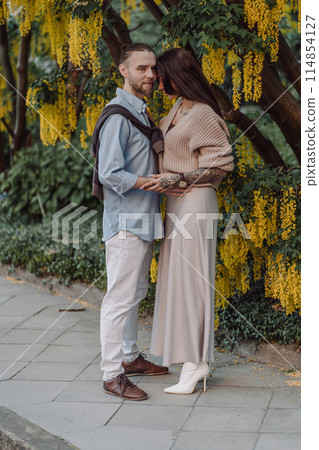 Amazing couple hugging under tree with yellow tree. Lovers couple hugging in a laburnum tree. Couple lovers hugging in the park. Girl with tattoos. A girl and a boy kiss. 114854127