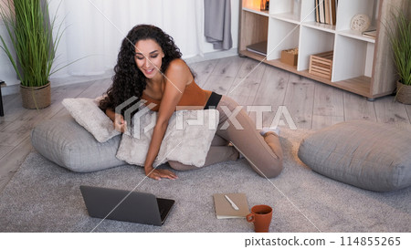 Video call. Online chat. Internet communication. Relaxed smiling woman speaking on laptop on floor at home living room interior with free space. 114855265