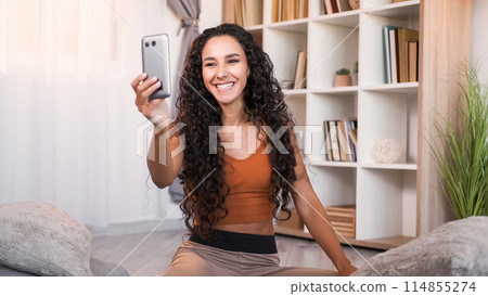 Online call. Virtual meeting. Internet conference. Happy smiling girl enjoying remote communication using mobile phone on floor at light modern home interior with copy space. 114855274