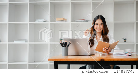A woman sitting at a desk with a laptop and a notebook. She is smiling and holding a pen. The scene suggests a productive and positive work environment 114857743