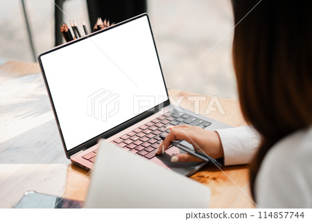 A woman is typing on a laptop with a blank screen. She is wearing a white shirt and has a pen in her hand. The scene suggests that she is working on a project or writing an email 114857744