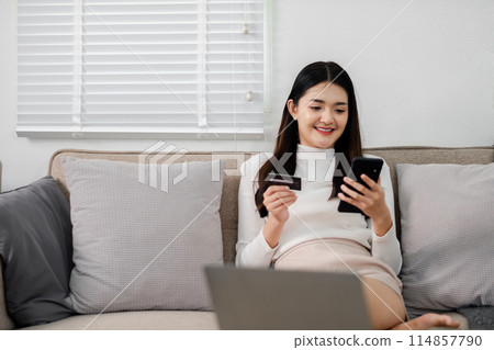 A woman is sitting on a couch and looking at her phone. She is holding a credit card and a laptop. Concept of relaxation and leisure, as the woman is using her phone and laptop to make a purchase 114857790