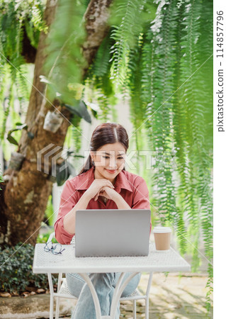 A woman is sitting at a table with a laptop in front of her. She is wearing a red shirt and has her hands crossed. The scene suggests a relaxed and comfortable atmosphere, possibly in a park or a cafe A woman is sitting at a table with a laptop in front of her. She is wearing a red shirt and has her hands crossed. The scene suggests a relaxed and comfortable atmosphere, possibly in a park or a cafe 114857796