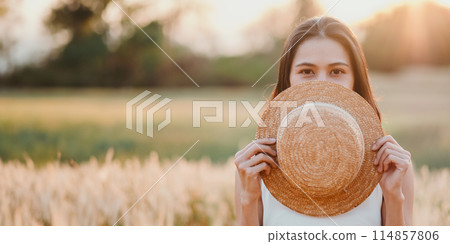 A woman is holding a straw hat and is hiding her face behind it. The image has a light and playful mood, as the woman is posing with the hat and making a silly face 114857806