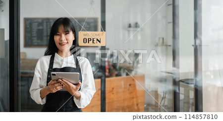 A woman wearing an apron stands in front of a sign that says "Welcome Open". She is holding a tablet in her hand A woman wearing an apron stands in front of a sign that says "Welcome Open". She is holding a tablet in her hand 114857810