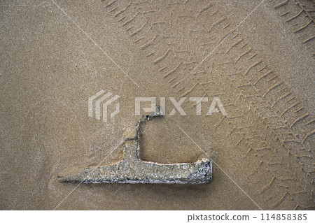 The metal part of ship covered with shells and buried in sand on seashore, with tire tracks nearby 114858385