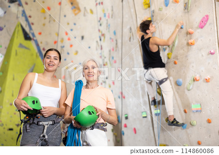 Two women of different ages standing with blue safety belts and helmet before practicing rock-climbing on climbing wall in adventure park 114860086