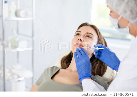 Beautician makeing cosmetic injection in the lips. Woman gets filler injection in her face. Beautician makeing cosmetic injection in the lips. Woman gets filler injection in her face. 114860571
