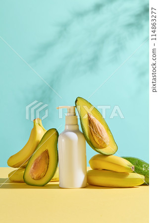A white bottle of cosmetic without label displayed in center of yellow table with fresh avocados and bananas leaning on, against blue color texture. Photo with front view for advertising 114863277