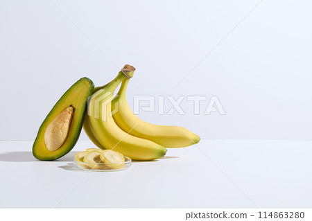 The photography of a banana bunch and a half of avocado leaning on each other, next to a glass dish of banana slices over white background. Front shot, blank space for showing product 114863280