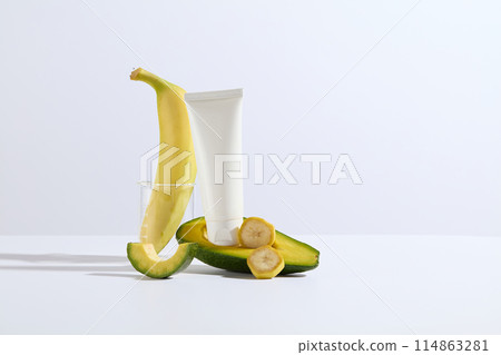The cosmetic advertising mock up photo on white background with banana and avocado ingredient, a white cosmetic tube without label displayed on a half of avocado decorated by some banana slices 114863281