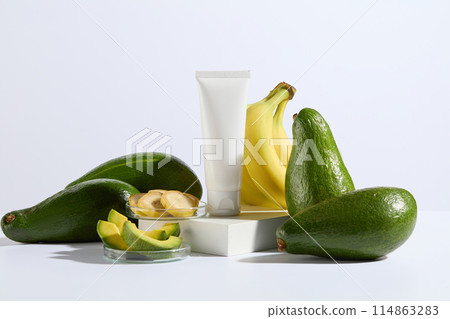Plenty fresh green avocados and yellow bananas decorated around a white platform containing a cosmetic tube without label above, glass dish of banana and avocado slices. Front view, space for design 114863283