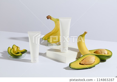White tone photograph of avocado and banana composition, two unlabeled cosmetic tubes are displayed on a table, 1 of which is placed on a white rectangular platform, fresh fruit arranged around White tone photograph of avocado and banana composition, two unlabeled cosmetic tubes are displayed on a table, 1 of which is placed on a white rectangular platform, fresh fruit arranged around 114863287