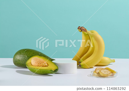 A small bunch of bananas and a few fresh avocados are scattered on white table around a circular empty platform in the middle, next to a glass dish with sliced banana against baby blue background A small bunch of bananas and a few fresh avocados are scattered on white table around a circular empty platform in the middle, next to a glass dish with sliced banana against baby blue background 114863292