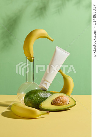Photo has creative layout of avocado and banana theme with frontal shot, a boiling flask with a banana on top, an unlabeled cosmetic tube placed on a avocado and leaning on a banana, space for design 114863317