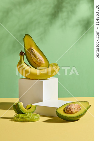 The yellow flatform containing a white platform with a banana bunch and a fresh avocado above, next to a blank podium, decorated with some fresh avocado in front over green background 114863320