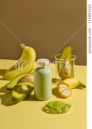 Vertical photo of high angle shot with a blank bottle of cosmetic displayed on yellow table, surrounded by a dish of chopped avocado and banana with some fresh avocado and banana, space for design 114863324
