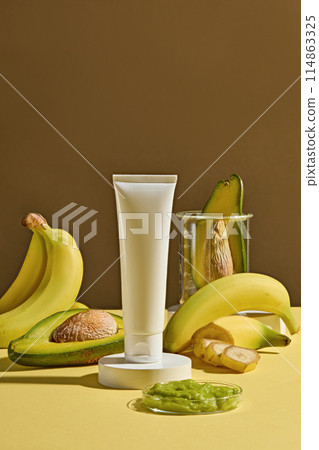 A small pedestal placed in the center of yellow table with an unbranded tube above, a glass dish of chopped avocado in front, fresh banana and avocado decorated in back, over brown background A small pedestal placed in the center of yellow table with an unbranded tube above, a glass dish of chopped avocado in front, fresh banana and avocado decorated in back, over brown background 114863325