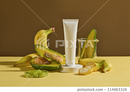 Yellow counter with fresh fruits ingredient above such as avocado and yellow banana, especially a blank label cosmetic tube for adding packaging of brand. Mock up for advertising with frontal shot 114863326