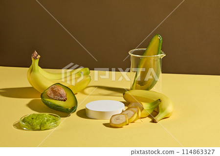 A empty small platform in white color displayed on yellow table top with a beaker containing a half avocado, chopped avocado, some bananas decorated around. Template for design with copy space 114863327