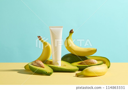 The cosmetic tube without label displayed on a half of fresh avocado, surrounded by plenty avocado and banana on yellow table top over blue background. Blank space for design, front view 114863331