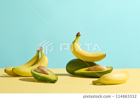 Fresh fruit ingredient as yellow banana and green avocado flat lay on yellow countertop against light blue background with many blank spaces for design. Photo was shot from front with copy space Fresh fruit ingredient as yellow banana and green avocado flat lay on yellow countertop against light blue background with many blank spaces for design. Photo was shot from front with copy space 114863332